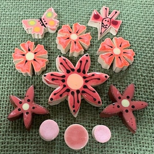 Peut inclure: Diverses pièces décoratives en céramique, comprenant des fleurs, des papillons et des étoiles. Les pièces sont principalement roses et rouges avec des accents noirs et jaunes, sur un fond vert texturé.