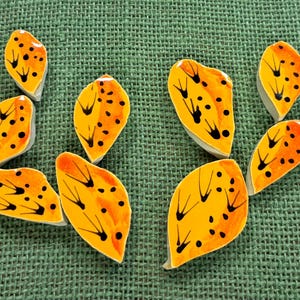 May include: Ceramic cactus pad ornaments in shades of yellow and orange, with black detailing. The pieces are arranged on a green, textured background. Each pad is approximately 5 cm long.