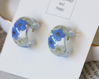 Puede incluir: Un par de pendientes de aro con una base de resina transparente y flores de nomeolvides azules prensadas en su interior. Los pendientes se muestran en una tarjeta blanca con el texto "Natural Power and Magic".