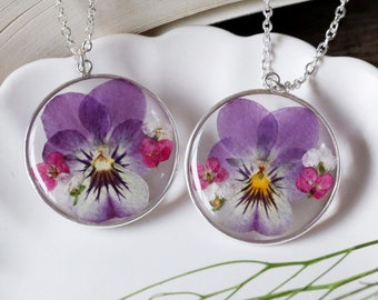 Puede incluir: Dos collares de plata con colgantes redondos que presentan pensamientos morados prensados ​​y flores blancas encapsuladas en resina transparente.