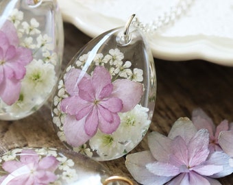 Puede incluir: Dos delicados collares con colgantes en forma de lágrima rellenos de flores rosadas y blancas prensadas. Los colgantes son transparentes y las flores son visibles en el interior. Los collares están sobre una superficie de madera con algunas flores sueltas.