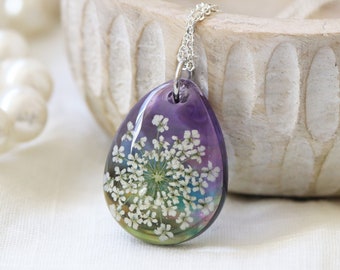 Puede incluir: Un collar de cadena de plata con un colgante en forma de lágrima. El colgante está lleno de resina transparente y contiene una flor blanca prensada. La resina tiene un brillo iridiscente púrpura, azul y verde.