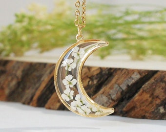 Puede incluir: Un collar de media luna dorado con flores blancas prensadas dentro de la resina. El collar está sobre una superficie de madera.