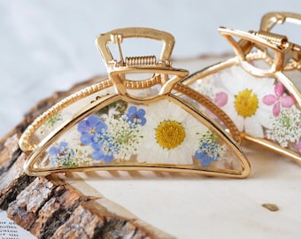 Puede incluir: Una pinza para el cabello de metal dorado con un diseño semicircular relleno de resina transparente. La resina contiene flores prensadas incrustadas, incluyendo margaritas blancas, pequeñas nomeolvides azules y follaje verde, con un resorte.