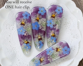 Puede incluir: Cuatro pinzas para el pelo de resina transparente con flores prensadas. Cada pinza presenta pensamientos morados, pequeñas flores azules y flores de relleno blancas. El texto "You will receive ONE hair clip" es visible.