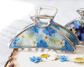 Puede incluir: Una pinza para el cabello semicircular con un marco de metal plateado. La superficie está rellena de resina transparente con flores prensadas incrustadas en tonos azules y blancos, incluyendo pétalos pequeños y delicados racimos florales.
