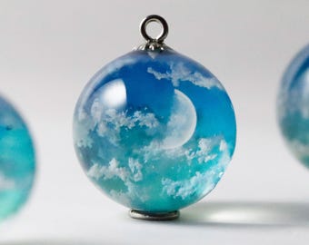 Puede incluir: Un colgante esférico con cuerpo de resina transparente y una escena de cielo incrustada. El diseño muestra un degradado de tonos azules, nubes blancas tenues y una luna creciente pálida. Un pequeño aro de metal plateado está en la parte superior.
