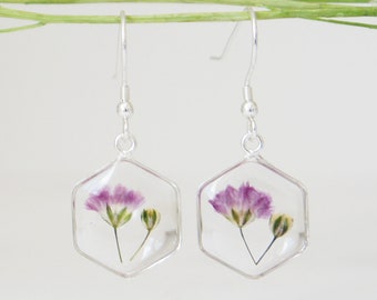Puede incluir: Un par de pendientes de plata con colgantes hexagonales de resina transparente con flores moradas prensadas.