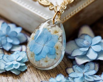 Puede incluir: Un colgante de resina en forma de lágrima que contiene pétalos de hortensia azul claro y pequeñas flores blancas. El colgante cuelga de una delicada cadena de tono dorado, con un acabado transparente y brillante que resalta los elementos botánicos.