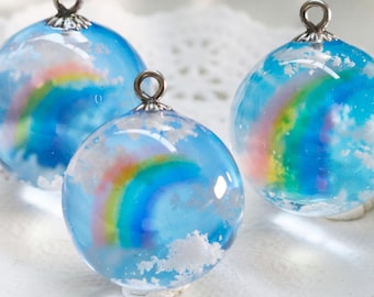 Puede incluir: Tres colgantes esféricos de resina con un fondo de cielo azul claro, un arcoíris multicolor vibrante y detalles de nubes blancas. Cada colgante está rematado con una pequeña anilla de metal plateado para colgar, con un acabado brillante.