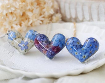 Puede incluir: Tres colgantes en forma de corazón con flores prensadas azules y blancas en su interior. Los colgantes están sobre una superficie blanca.