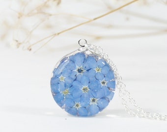 May include: A round pendant necklace featuring a cluster of small, light blue flowers encased in clear resin. The pendant hangs from a delicate silver chain. The background is white with a hint of dried flowers.