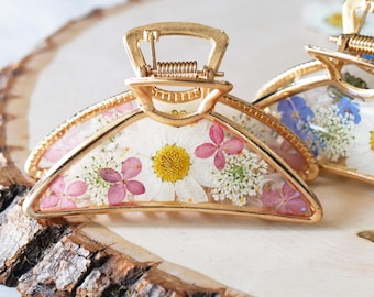 Puede incluir: Una pinza para el cabello de metal dorado con un diseño semicircular relleno de resina transparente. Incrustadas en la resina hay flores de margarita blancas prensadas, pequeños pétalos de hortensia rosa y delicadas flores blancas.