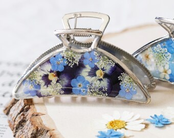 Puede incluir: Una pinza para el cabello de metal plateado con un diseño semicircular relleno de resina transparente. La resina contiene flores prensadas incrustadas, incluyendo pensamientos de color púrpura oscuro, pequeñas nomeolvides azul claro y flores blancas.