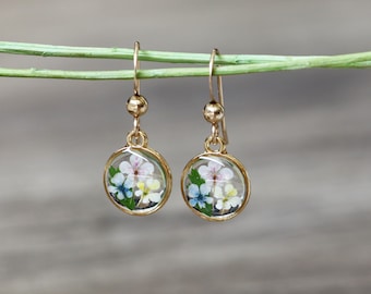 Puede incluir: Un par de pendientes de aro dorados con colgantes redondos de resina transparente. Cada colgante presenta un grupo de flores prensadas en tonos de blanco, rosa, azul y amarillo.
