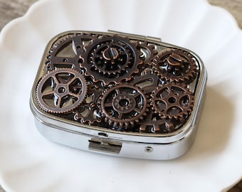 Puede incluir: Una caja de píldoras de plata con un diseño decorativo de engranajes de cobre. La caja tiene una tapa con bisagras y un cierre.