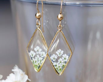 Puede incluir: Un par de pendientes colgantes de tono dorado con colgantes de resina transparente en forma de diamante. Cada colgante contiene flores blancas prensadas con tallos verdes visibles dentro del material transparente.