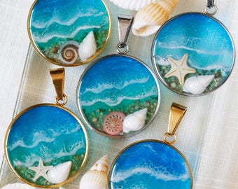 Puede incluir: Cinco colgantes circulares con una escena oceánica de resina con agua azul y turquesa, olas de espuma blanca y orillas arenosas. Cada colgante incluye pequeñas conchas marinas o una estrella de mar incrustadas en un marco metálico.