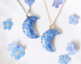 Puede incluir: Dos collares de media luna llenos de pequeñas flores azules. Uno tiene una cadena plateada, el otro una cadena dorada. Varias flores azules sueltas están esparcidas alrededor de los collares sobre un fondo blanco.
