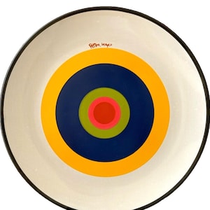 Könnte beinhalten: Ein weißer Keramikteller mit schwarzem Rand zeigt ein konzentrisches Kreisdesign in Gelb, Blau, Grün, Rosa und Rot. Der Teller ist mit "Peter Maxo" signiert.