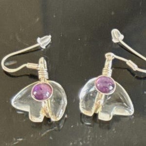 May include: A pair of dangle earrings featuring clear, carved bear-shaped pendants made of translucent stone. Each bear is accented with a small, round, purple amethyst cabochon set at the top. The earrings hang from silver-toned metal hooks.