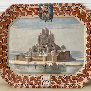 May include: A ceramic tile with a painted image of Mont Saint-Michel, a historic abbey located on a tidal island in Normandy, France. The tile is decorated with a brown and white border and a small ceramic angel in the top corner. The tile reads "Mont St Michel".