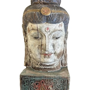 Puede incluir: Una escultura de madera de una cabeza de Buda con una corona azul y dorada, ojos cerrados y un punto rojo en la frente. La cabeza está montada sobre una base de madera con un diseño de flor de loto tallado en verde, rojo y dorado.