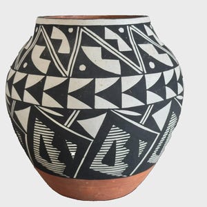 May include: A rounded ceramic vase featuring a wide opening and a terracotta-colored base. The upper body is decorated with a bold black and white geometric pattern consisting of triangles, lines, and angular shapes arranged in horizontal bands.