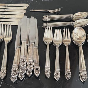 May include: A collection of silver-toned flatware including multiple knives, forks, and spoons. Each piece features a decorative, ornate handle with intricate scrollwork at the base. The set includes various sizes of forks and spoons arranged in groups.