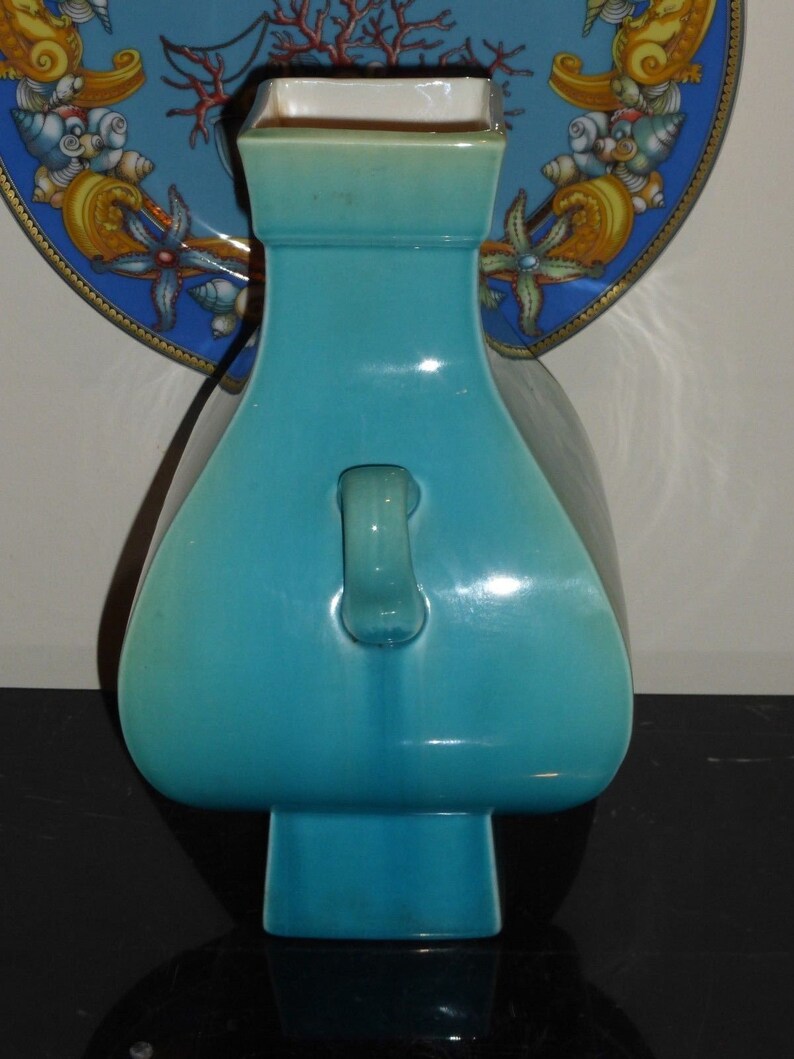 Pode incluir: Um vaso de cer&acirc;mica turquesa com uma base quadrada e uma al&ccedil;a no lado. O vaso tem um acabamento liso e brilhante.