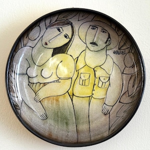 May include: Round ceramic plate with a black rim, featuring a hand-painted illustration of a man and woman. The man wears a yellow shirt and hat, while the woman is in a yellow dress. The plate is signed "MANUEL".