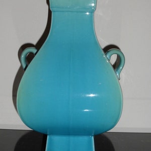 Pode incluir: Um vaso de cer&acirc;mica turquesa com duas al&ccedil;as e uma base quadrada. O vaso tem um acabamento liso e brilhante.