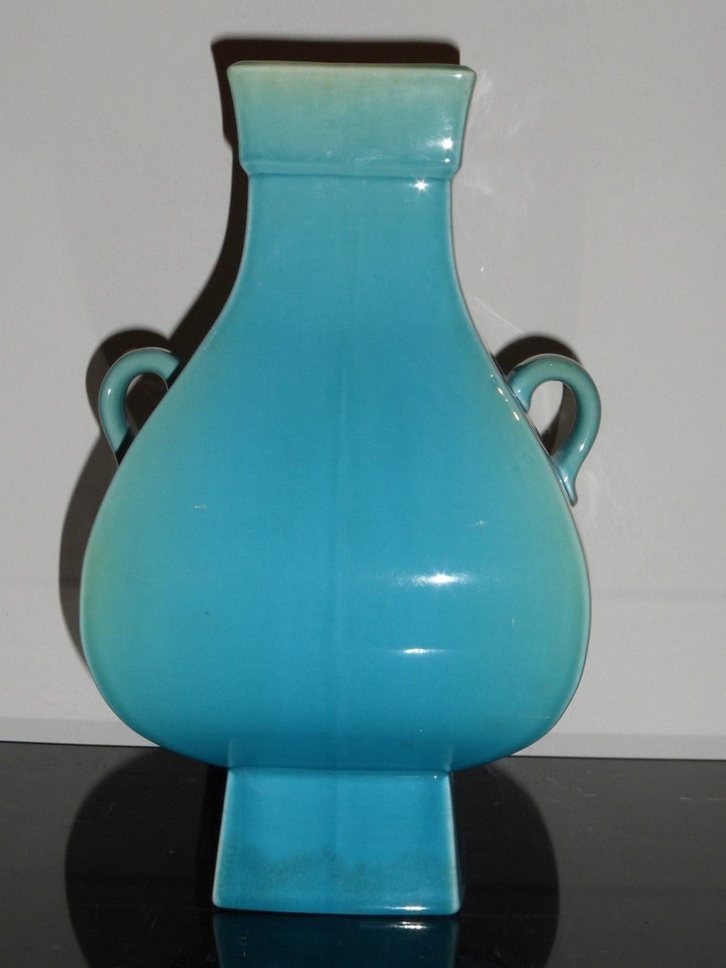 Pode incluir: Um vaso de cer&acirc;mica turquesa com duas al&ccedil;as e uma base quadrada. O vaso tem um acabamento liso e brilhante.