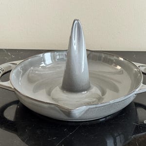 Può includere: Un arrostitore per pollo in ceramica grigia di forma rotonda con un cono centrale verticale per la cottura in verticale. Il piatto presenta due manici laterali, ciascuno impresso con il marchio STAUB, e una finitura liscia e lucida sfumata.