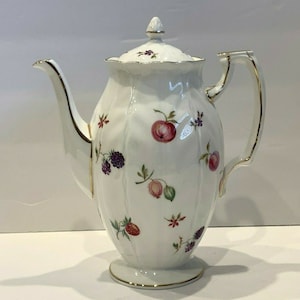 Puede incluir: Cafetera de porcelana blanca con tapa, asa y boquilla. La cafetera está decorada con diseños florales y frutales coloridos y ribetes dorados. La cafetera tiene una capacidad aproximada de 1,2 litros.