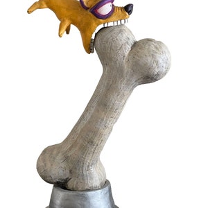 May include: A yellow and purple dog sculpture with a wild expression is biting down on a large bone. The bone is on a silver base.