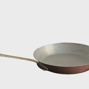 May include: A copper-bottomed frying pan with a long, brass-colored handle. The interior of the pan is silver-colored. The pan is designed for cooking and is suitable for use in a kitchen.