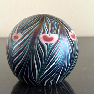 Può includere: Un fermacarte sferico in vetro con un motivo a piume di pavone. I colori dominanti sono blu e verde acqua, con accenti marroni e rosa. Il vetro ha una finitura iridescente che riflette la luce. Circa 8 cm di diametro.