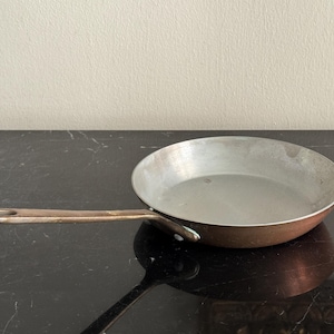 May include: A copper-bottomed frying pan with a long, riveted handle. The interior is silver-colored, and the pan sits on a dark surface. The pan is designed for cooking and is a kitchen essential.