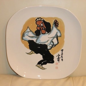 Könnte beinhalten: Ein weißer Keramikteller mit einer handgemalten Illustration einer Comicfigur in einem weißen Gewand und schwarzen Hosen. Die Figur befindet sich in einer Kampfpose vor einem gelben Hintergrund. Der Teller ist mit schwarzer Tinte mit dem Text "郭子明" und einem roten Siegel signiert.