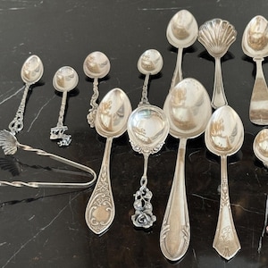 May include: An assortment of antique silver spoons and serving tongs. The spoons feature various handle designs, including floral, shell, and ornate patterns. The tongs have a twisted handle and shell-shaped ends. The spoons are arranged on a dark surface.