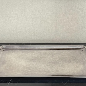 May include: A rectangular silver serving tray with decorative handles. The tray has a simple, elegant design and is likely made of metal. The handles feature a scrolled design. The tray is placed on a dark surface.