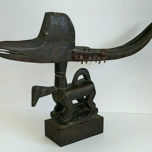 Puede incluir: Una escultura de madera oscura de un pájaro estilizado con un pico largo y curvo y una cola levantada. La escultura está decorada con cuentas rojas y se apoya sobre una base de madera.