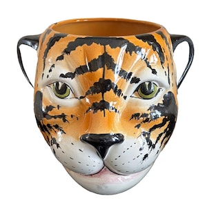 Pode incluir: Um vaso de cerâmica em forma de cabeça de tigre. O vaso é pintado em tons de laranja, preto e branco para se assemelhar à pelagem de um tigre. O vaso tem duas alças nas laterais.