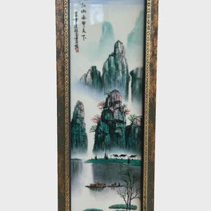 May include: A framed Asian landscape painting featuring mountains, water, and small boats. The artwork is in shades of green, blue, and brown, with a gold-colored frame. The painting includes Chinese characters.