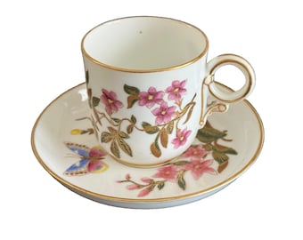 May include: A white teacup and saucer set with gold trim. The cup and saucer feature pink floral and butterfly designs. The cup's handle has an ornate design. A classic tea set.