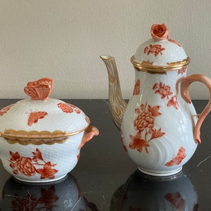 Puede incluir: Un juego de té de porcelana blanca con un patrón floral en tonos naranja y dorado. El juego incluye una tetera y un azucarero con tapa. Ambas piezas presentan delicados detalles de mariposa.