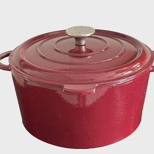 Puede incluir: Una olla holandesa de hierro fundido de color burdeos con tapa y dos asas laterales. La tapa tiene un pomo plateado. La olla redonda tiene un acabado brillante, ideal para cocinar guisos, sopas y estofados.