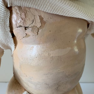 May include: A vintage porcelain doll with a white knitted shirt. The doll's body is made of porcelain and has a cracked and chipped surface. The doll's face is not visible in the image.