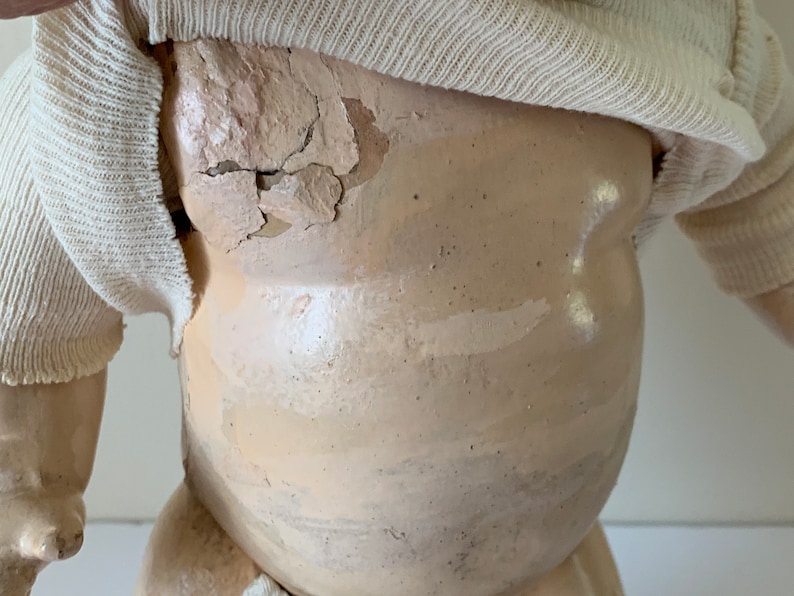 May include: A vintage porcelain doll with a white knitted shirt. The doll's body is made of porcelain and has a cracked and chipped surface. The doll's face is not visible in the image.